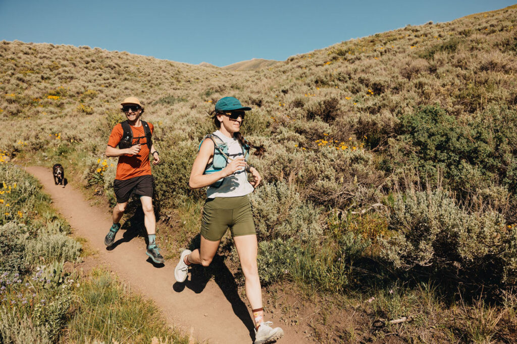Two people trail running on a dirt path through a hilly, grassy landscape under a clear blue sky, wearing athletic gear and sunglasses, with a dog running behind them.