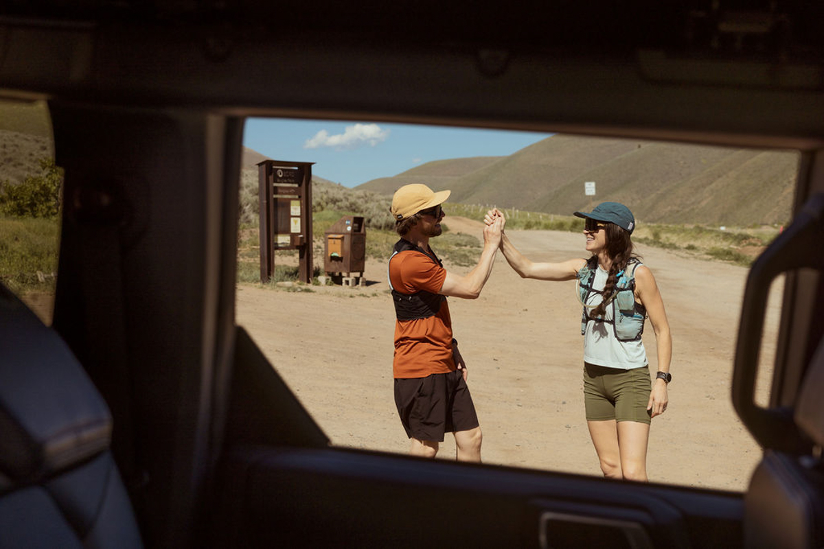 Two hikers wearing hats and backpacks high-five each other in a sunny, outdoor parking area, viewed through a car window, with hills and a trail sign in the background.