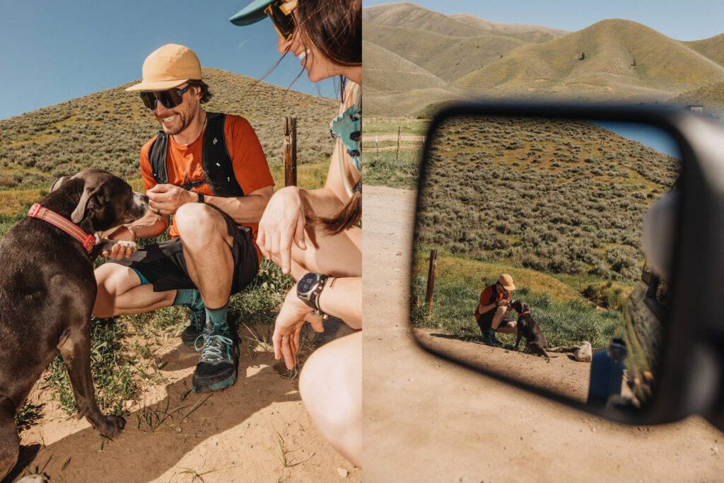 Two people sit on a dirt path in grassy hills, smiling and feeding a black dog. The right side shows their reflection in a car mirror with the same scene and hills in the background under a sunny sky.
