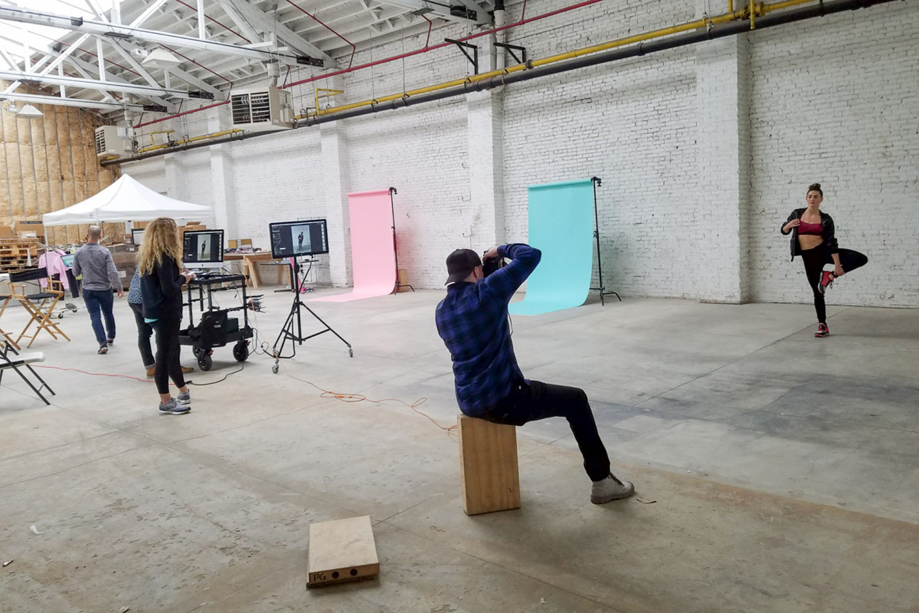 A photographer captures a woman posing before colored backdrops in a spacious, industrial-style studio, while crew members with equipment and monitors prepare for seamless post production services.