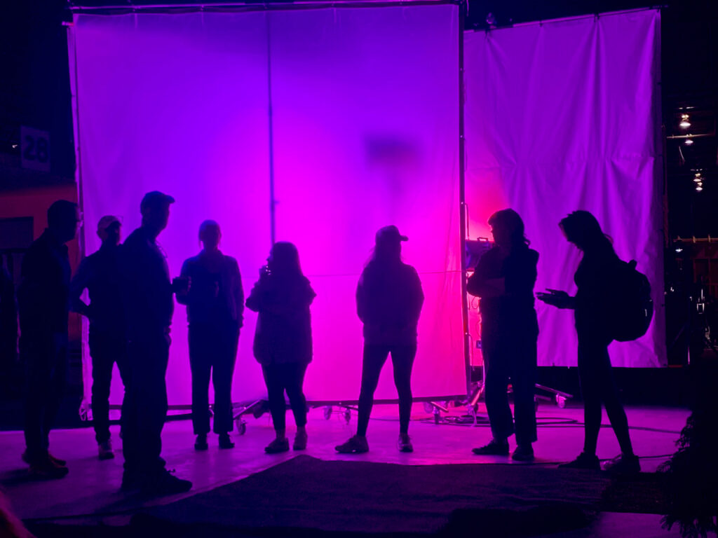 Silhouettes of seven people stand in front of a large, brightly lit purple and pink backdrop, casting dramatic shadows. The creative, backstage atmosphere hints at the artistry behind post production services. Some chat or use their phones.