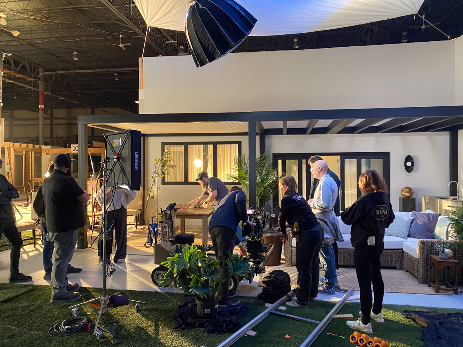 A film crew sets up lighting and equipment around actors in a staged modern living room set, with cameras, track, and props visible on an indoor soundstage—capturing footage ready for expert post production services.