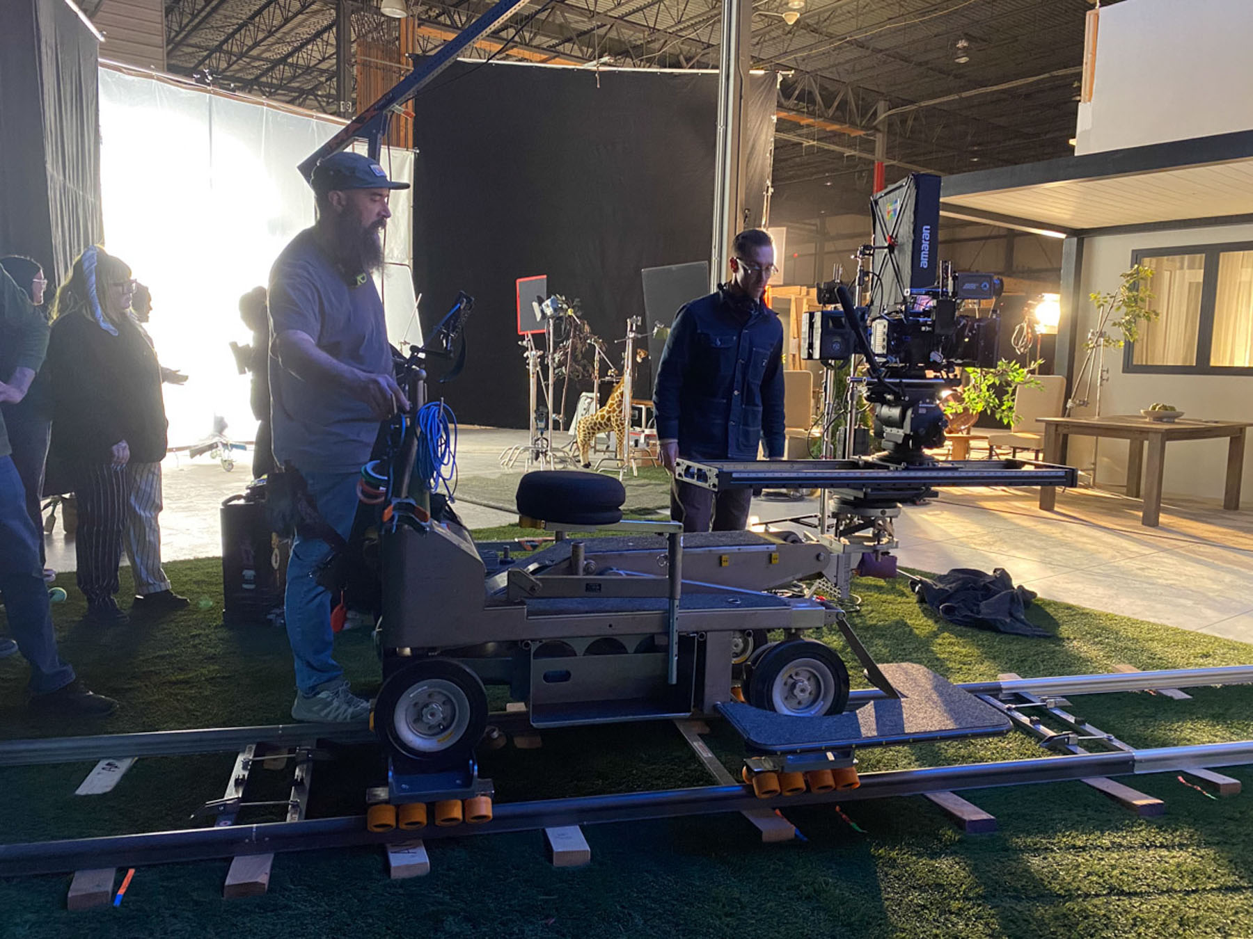 Film crew members operate a camera dolly on tracks inside a studio set, surrounded by lighting equipment and props. The environment suggests a professional movie or TV production, with post production services enhancing the final results.