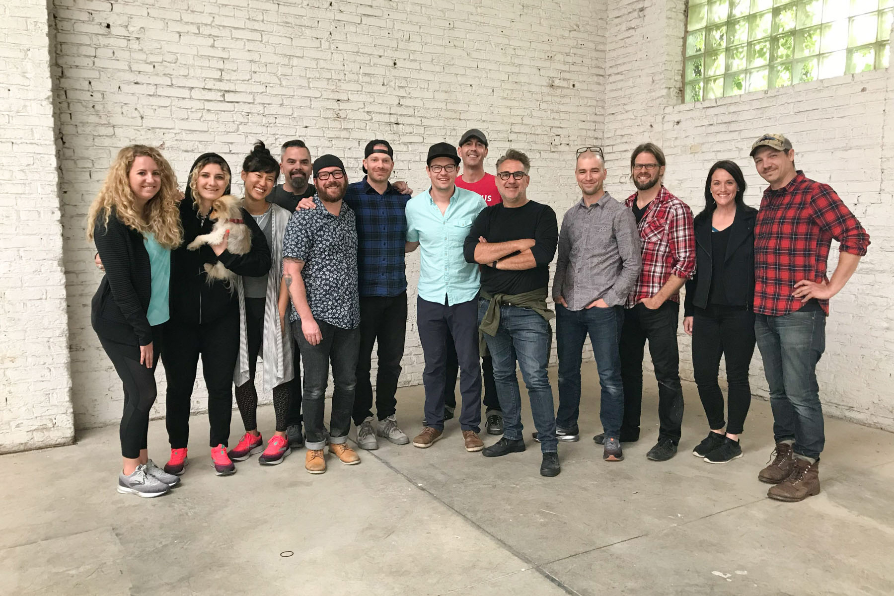 A group of thirteen people, some smiling and some with neutral expressions, stand together indoors against a white brick wall. One person holds a small dog. The casually dressed group appears to be part of a post production services team.