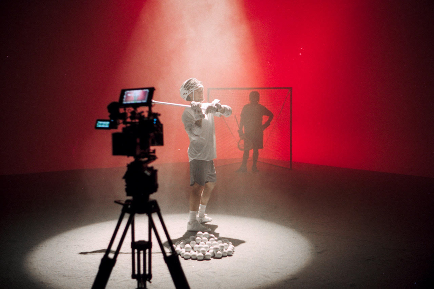 A person stands in a spotlight holding a baseball bat, surrounded by baseballs, with a camera filming them—perfect for showcasing post production services. Another person stands near a net in the dramatic red-lit backdrop.