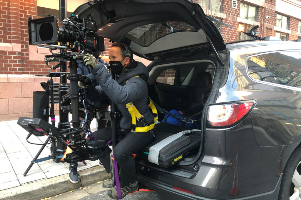 A masked cameraman wearing a harness operates a large camera rig from the open trunk of an SUV parked on a city street, with brick buildings in the background—capturing footage for post production services.