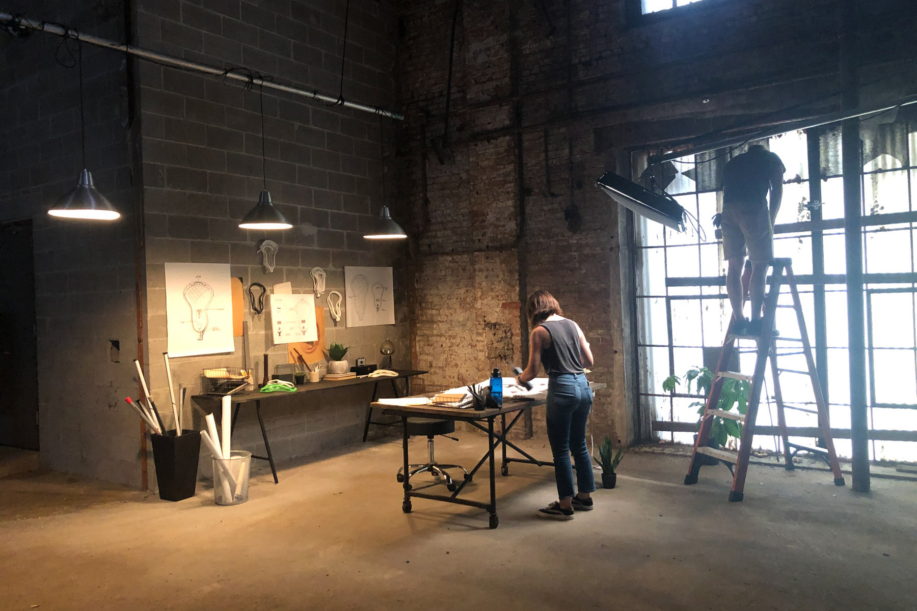 A person stands at a table working with papers while another on a ladder adjusts something near a large window in an industrial-style room, suggesting a creative space where post production services might be utilized amid exposed brick and sketches.