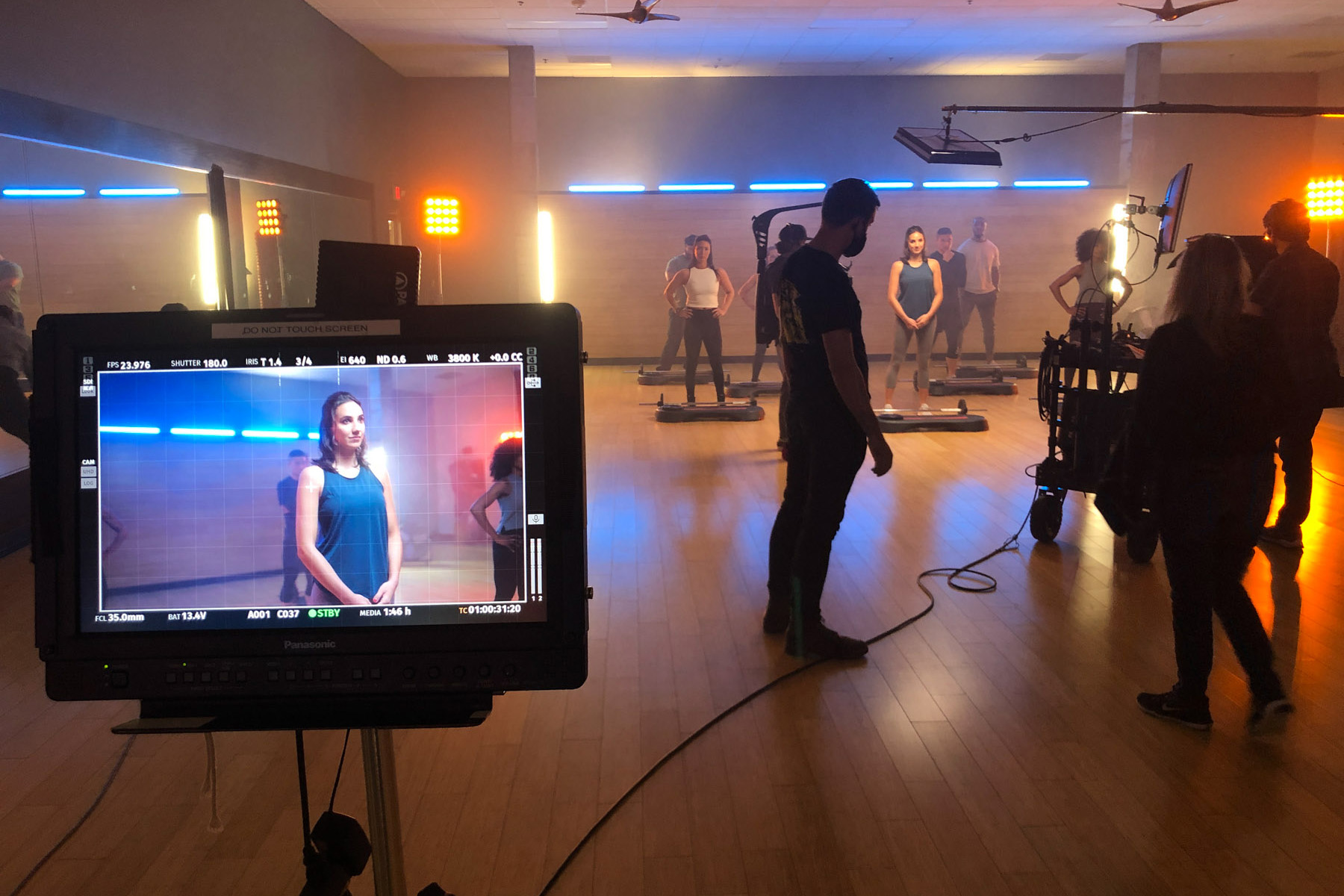 A film crew shoots a scene in a studio with actors standing in a row, while a monitor in the foreground displays a close-up of a woman on set. The room is lit with orange and blue lights, showcasing the importance of post production services.