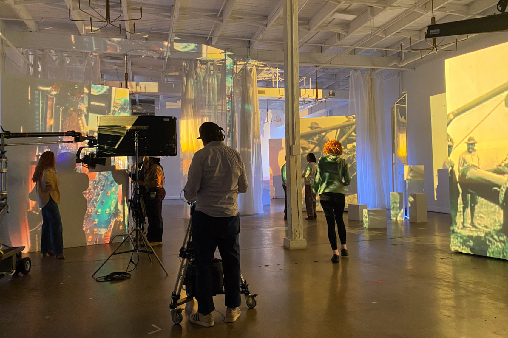 Several people are inside an art gallery or studio with large video projections on the walls, including vintage scenes. A person operates a camera, while others observe—possibly exploring post production services to enhance the experience.