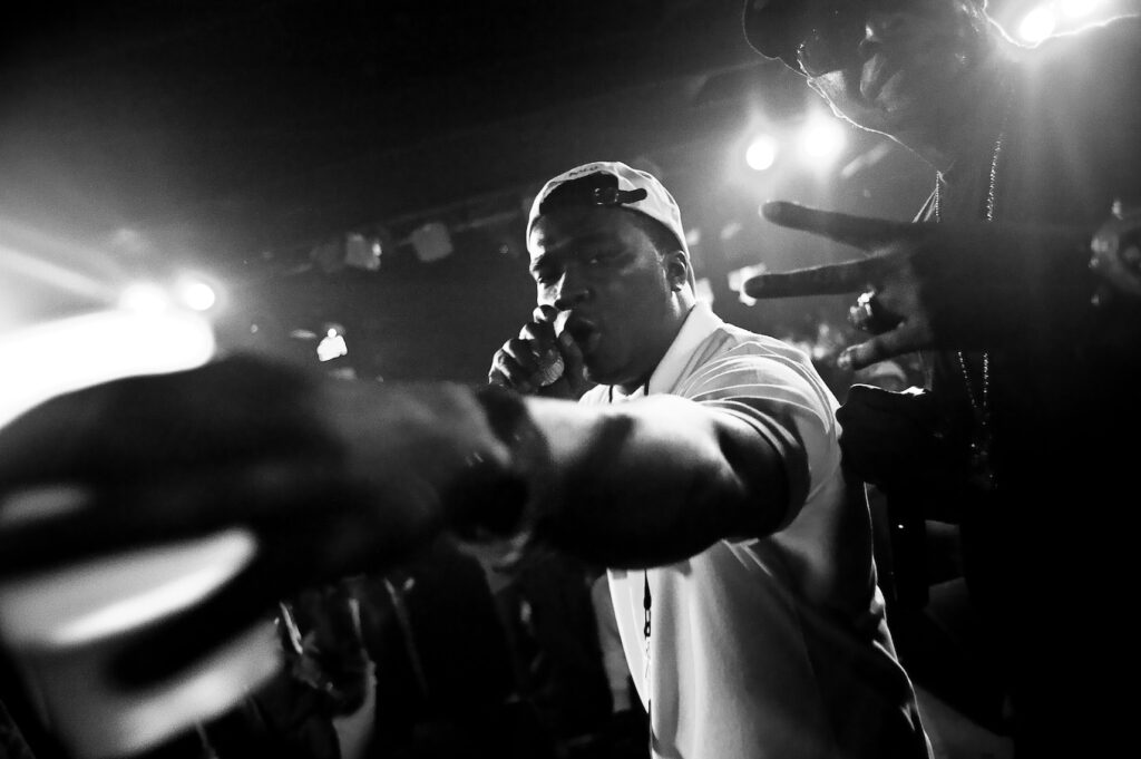 A black and white photo of a performer holding a mic and reaching toward the camera, wearing a backwards cap, with another person beside him flashing a peace sign in a lively, dimly lit venue.