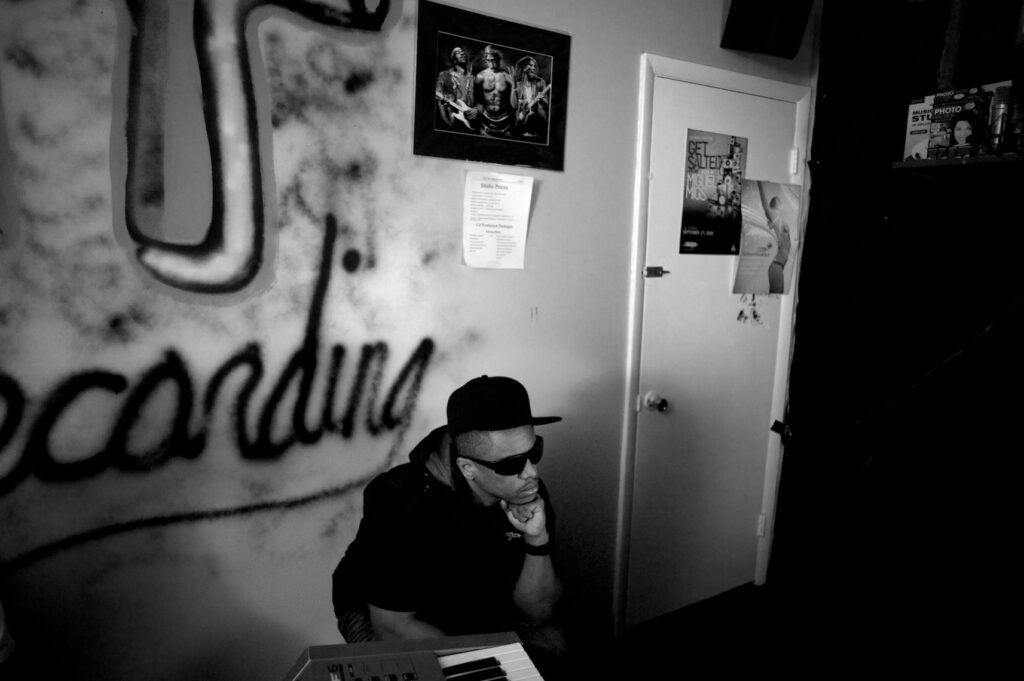 A man wearing sunglasses sits pensively in a dimly lit recording studio, with graffiti-style wall art, a keyboard, posters, and framed photos visible in the background.