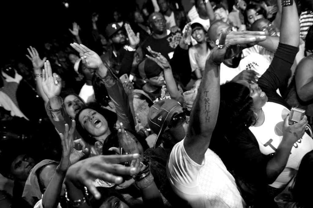 A lively crowd at a concert raises their arms and cheers, expressing excitement and energy. Some people hold phones to capture the moment. The scene is in black and white.