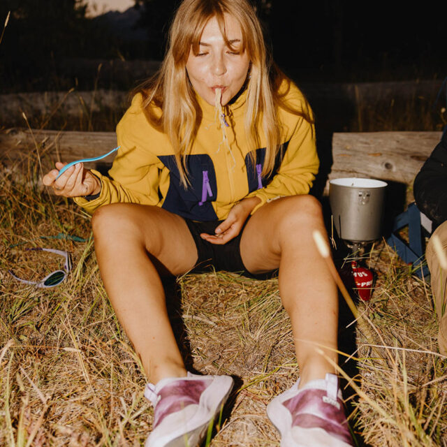 A woman sitting on grass spills noodles from her mouth while eating with a fork. She wears a yellow jacket and sneakers, with a cooking pot and camping stove beside her.