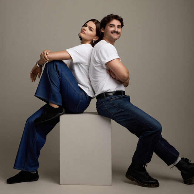 A woman and a man sit back-to-back on a cube, both wearing white t-shirts, blue jeans, and black shoes, posing confidently against a plain gray background.