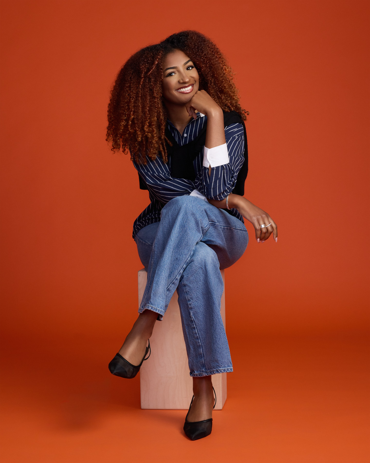 A woman with curly hair, wearing a striped shirt, jeans, and black heels, sits on a wooden block against an orange background, smiling with one hand resting on her chin.