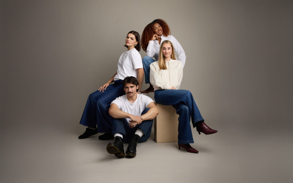 Four people pose together in a studio with neutral background. Three women sit and stand around a wooden block, while a man sits cross-legged on the floor in front. All wear casual tops and blue jeans.