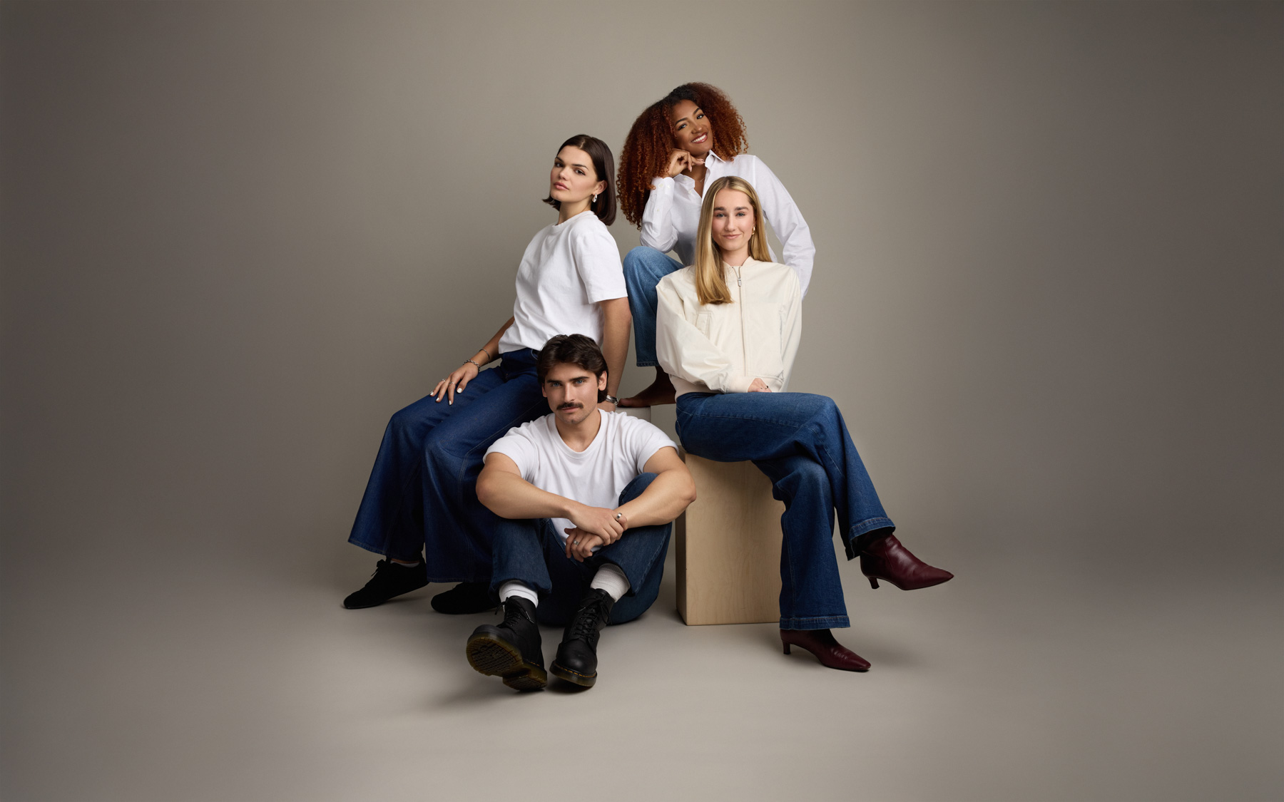 Four people pose together in a studio with neutral background. Three women sit and stand around a wooden block, while a man sits cross-legged on the floor in front. All wear casual tops and blue jeans.