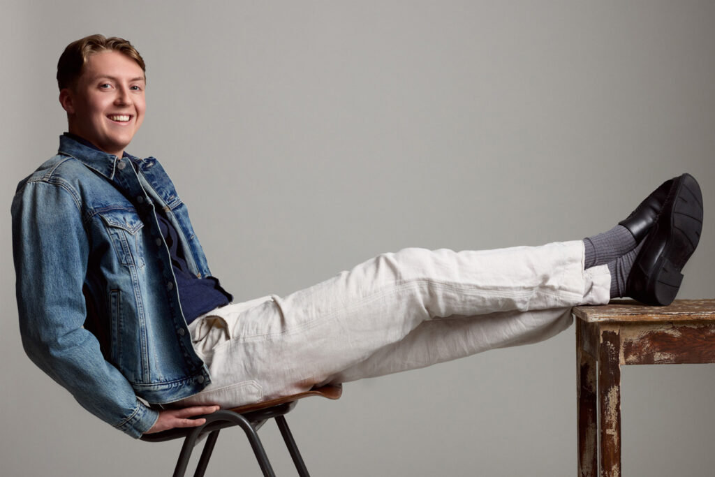 A smiling young man in a denim jacket and white pants reclines on a chair with his legs stretched out onto a wooden table, posing against a plain light gray background.