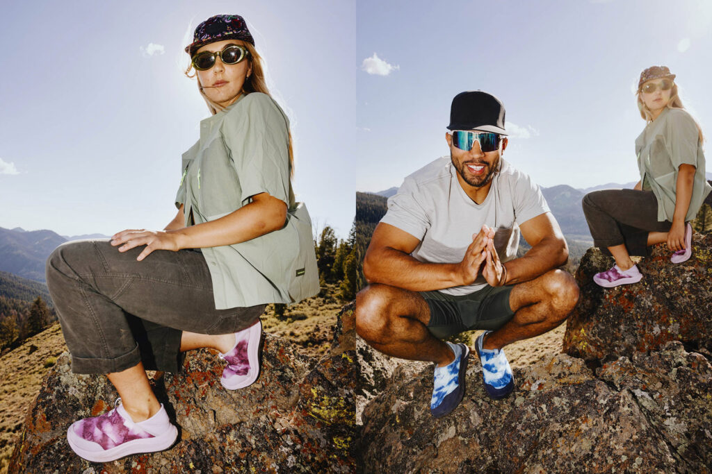 Two people wearing casual outdoor clothing and colorful slip-on shoes sit on rocky terrain with mountains in the background, enjoying a sunny day. The woman wears purple shoes; the man wears blue shoes and sunglasses.