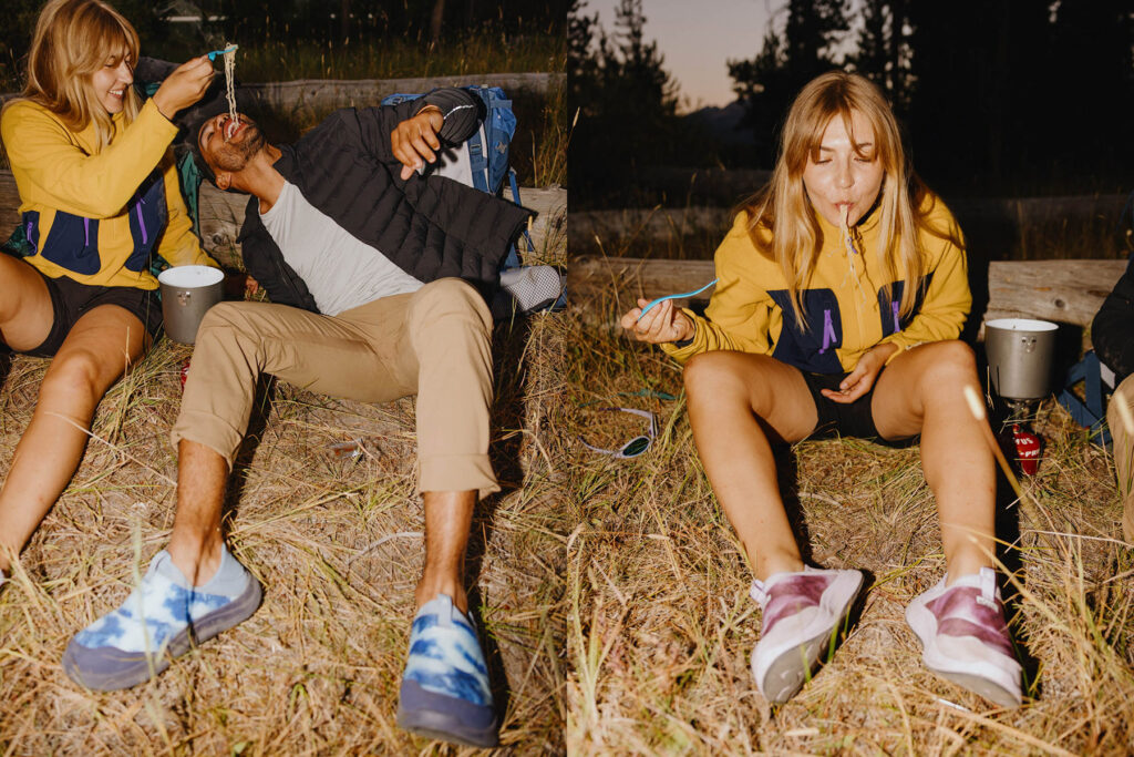 Two people sit outdoors at dusk, one woman in a yellow hoodie playfully feeds a man lying back in casual clothes; in the second frame, she eats with a spoon, surrounded by camping gear and grass.