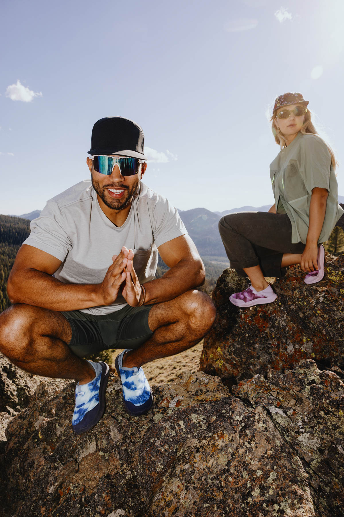 A man squats on a rocky mountain, smiling and wearing sunglasses, a black cap, and blue shoes. A woman sits nearby, also wearing sunglasses, a cap, and pink shoes. The sky is clear with mountains in the background.