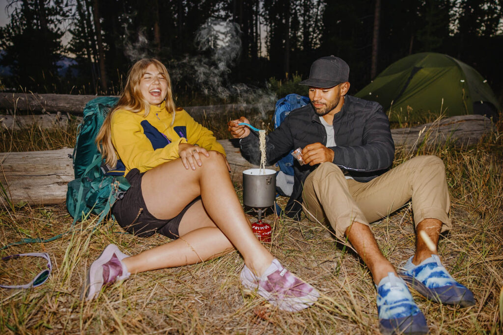 Two people sit on grass near a tent in the woods, laughing and cooking noodles on a portable stove. They wear casual outdoor clothing and backpacks, enjoying a relaxed camping moment in the evening.