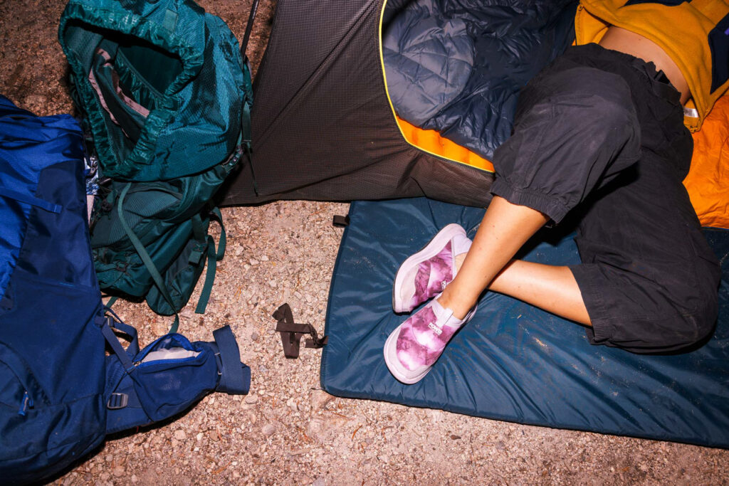A person wearing pink slippers and black pants lies on a sleeping pad at a campsite beside a tent, with green and blue backpacks resting on the rocky ground nearby.