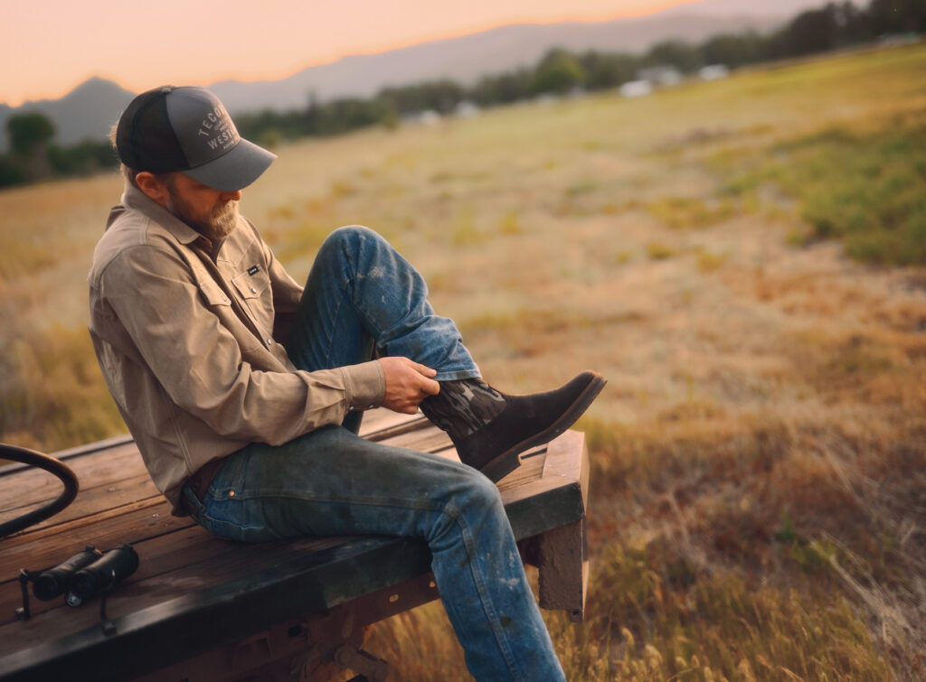 A man in classic workwear—a cap, beige shirt, and blue jeans—sits on a wooden surface outdoors at sunset, pulling on dark boots. Surrounded by tall grass with mountains in the background, he prepares for a hunting adventure.