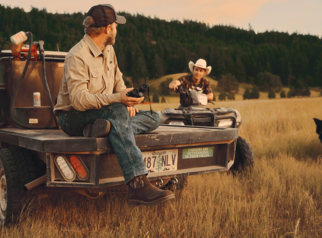 A man in boots sits on the back of a truck holding binoculars, watching another man in a cowboy hat on an ATV in a grassy field surrounded by trees at sunset—a perfect scene for hunting enthusiasts.