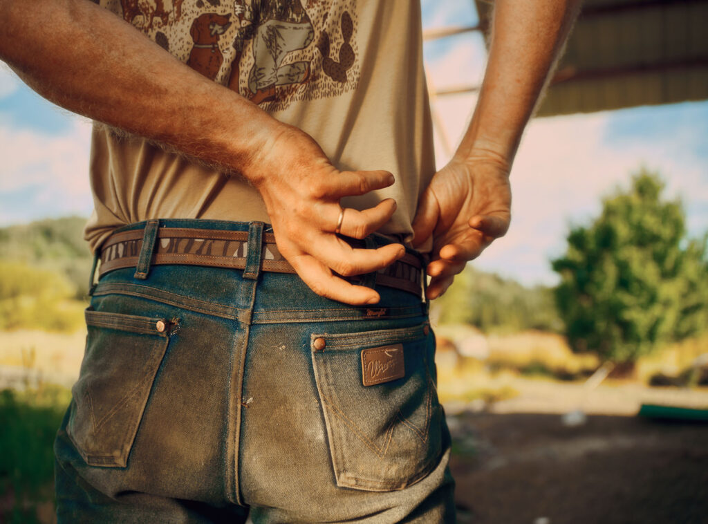 A person wearing jeans and a graphic t-shirt adjusts their waistband outdoors, with sunlight highlighting their hands and back. A field and tree are blurred in the background.
