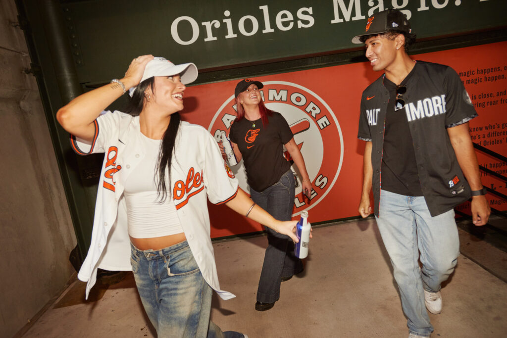 Three people wearing Baltimore Orioles jerseys and jeans smile and walk together in a stadium, with an Orioles logo and “Orioles Magic” sign in the background.