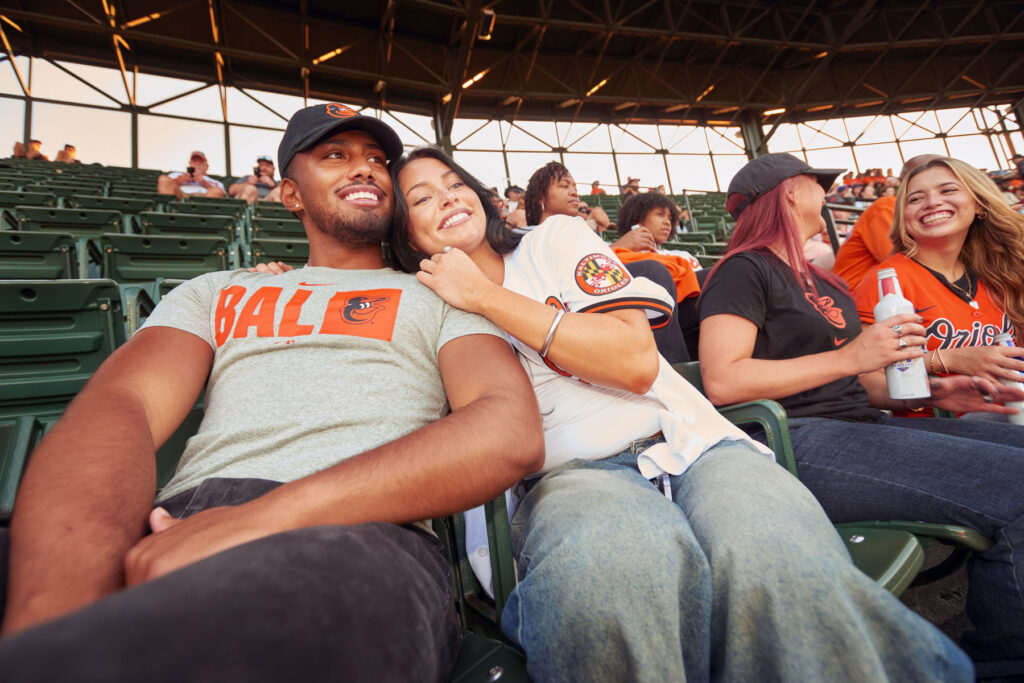 A group of young adults in Orioles gear smile and relax in stadium seats during a baseball game. Two people in the front row pose happily together, while others chat and hold drinks in the background.
