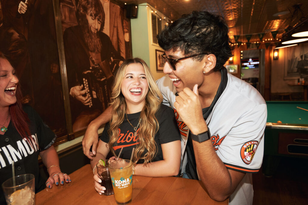 Three friends wearing baseball jerseys laugh together at a bar table. One woman holds a drink with a lime slice. A billiards table and musical murals are visible in the warmly lit background.