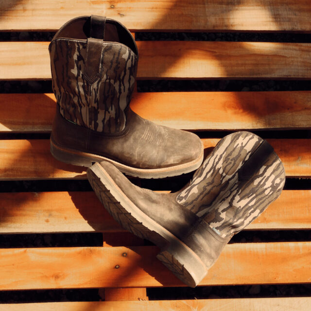 A pair of brown Tecovas cowboy boots with a camouflage pattern from Mossy Oak on the shafts are placed on a wooden surface with sunlight casting shadows across them.