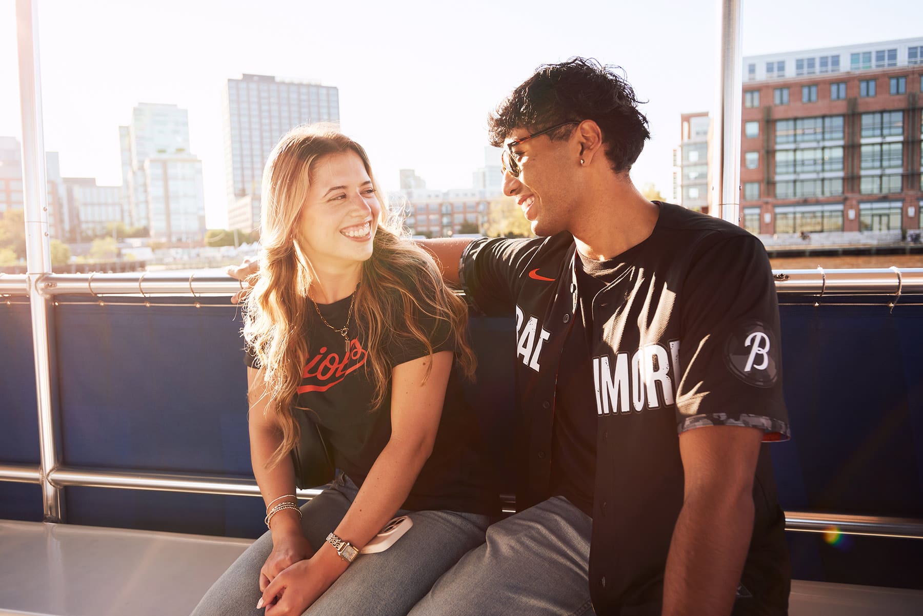 A young woman and man sit together on a bench outdoors, smiling at each other. The sun is shining, and modern buildings are visible in the background. They both wear casual sports jerseys.
