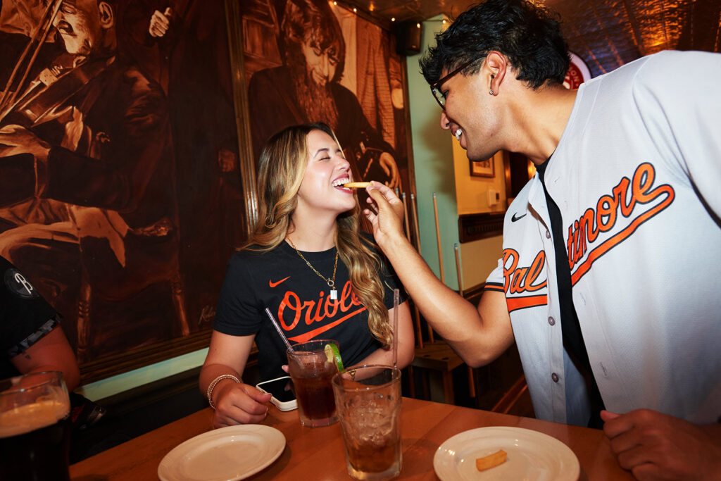 A smiling man in a Baltimore Orioles jersey feeds a French fry to a laughing woman, also in an Orioles shirt, at a restaurant table with drinks and empty plates. Large jazz-themed paintings are visible in the background.