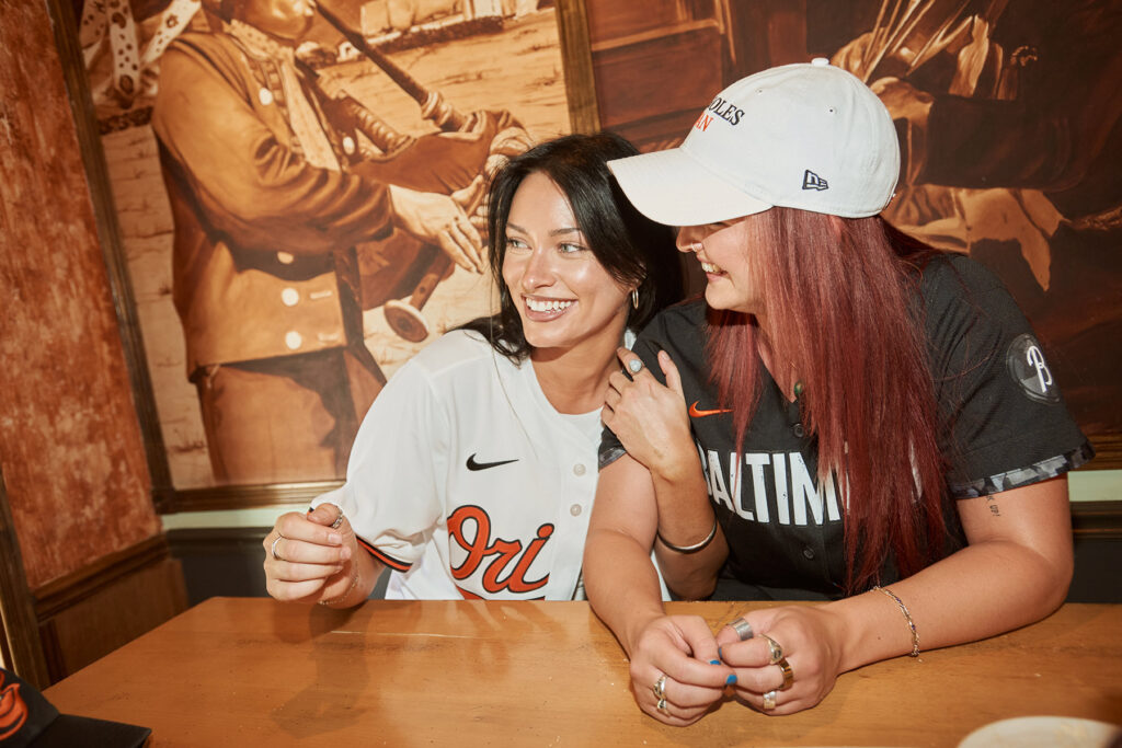 Two women in Baltimore Orioles baseball jerseys sit at a wooden table, smiling and laughing together; one wears a white cap, and they are in front of a wall with large sepia-toned historical images.