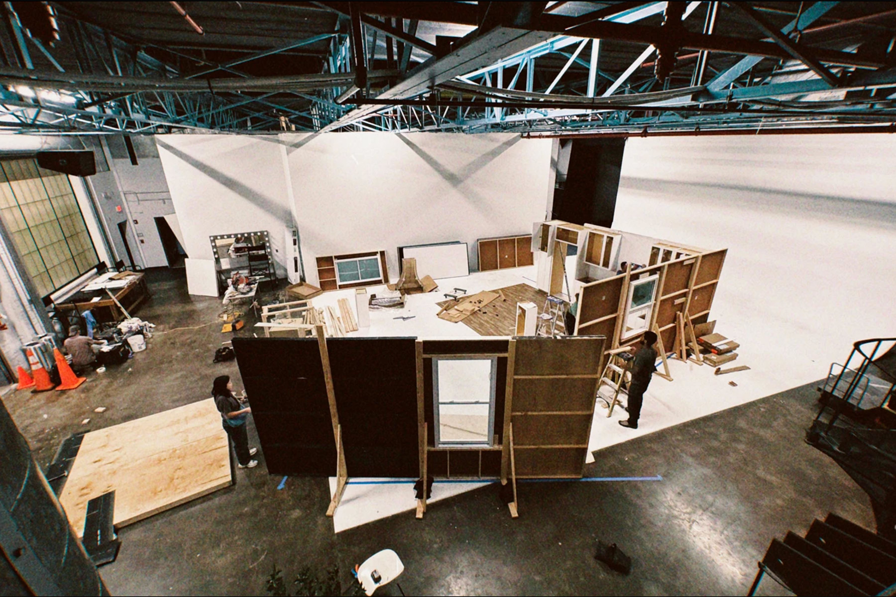 A film set under construction in a large studio, with people assembling modular wooden walls and various building materials scattered on the floor. Exposed ceiling beams and studio lights are visible above.
