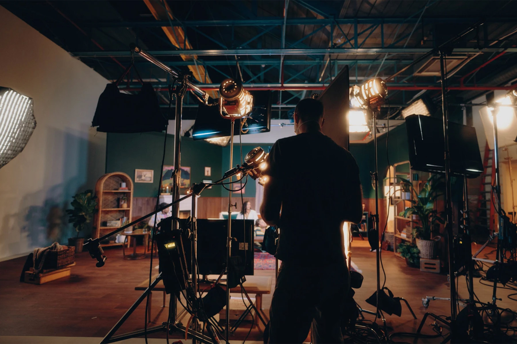 A person stands on a film set surrounded by cameras, lights, and equipment, preparing for a shoot in a spacious studio with furniture and plants in the background.