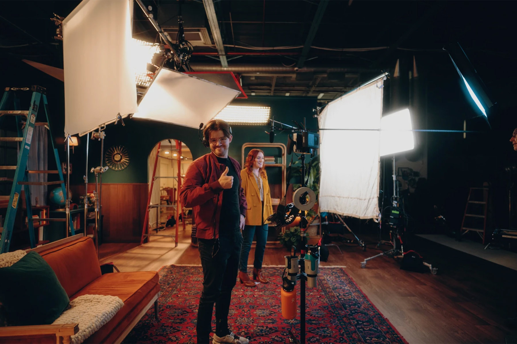 A man stands on a film set giving a thumbs-up, with a woman in the background. The set is filled with cameras, lights, and filming equipment, and features a couch and patterned rug. The atmosphere is creative and professional.