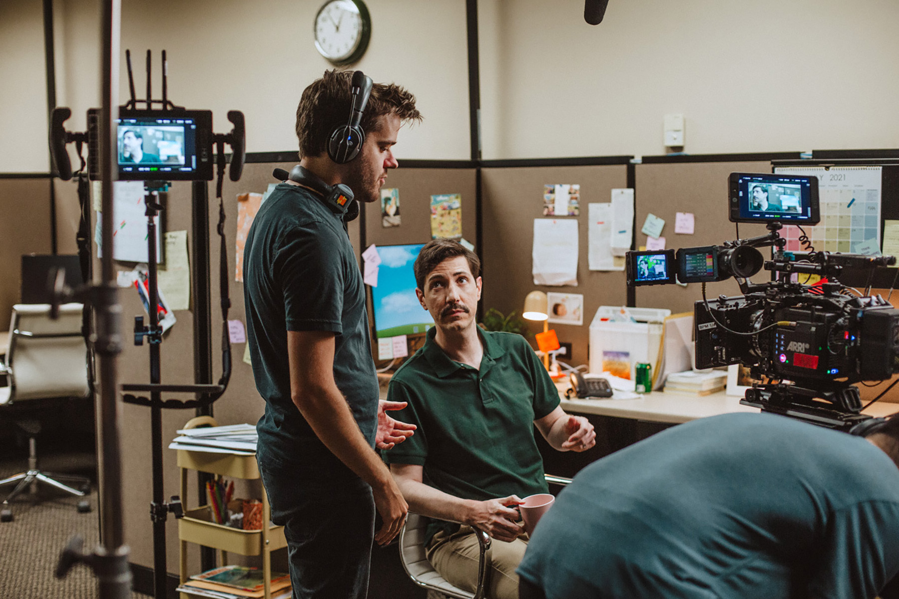 Two men talk on a film set in an office, surrounded by cameras, monitors, and lighting equipment. One is seated at a desk holding a cup, while the other stands nearby wearing headphones.