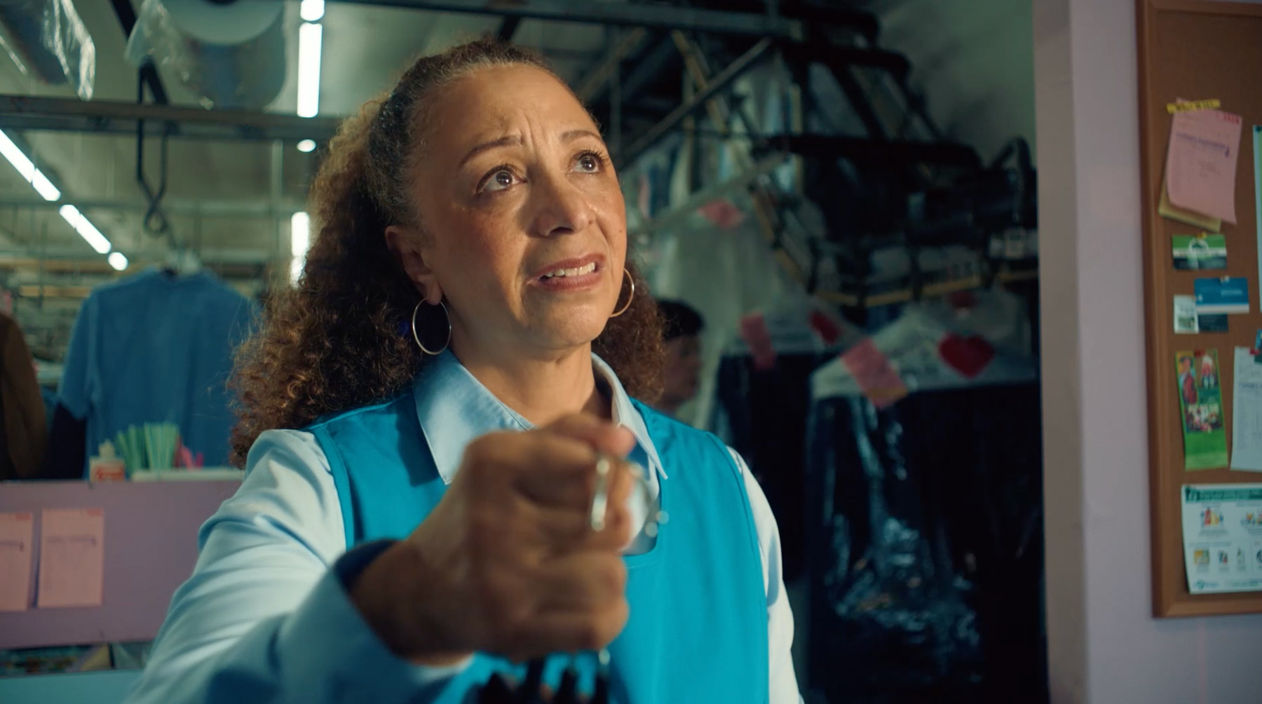 A woman in a blue uniform stands in a dry cleaning shop, holding out a set of keys with a concerned expression. She has curly hair pulled back and wears hoop earrings. Garments hang in plastic behind her.