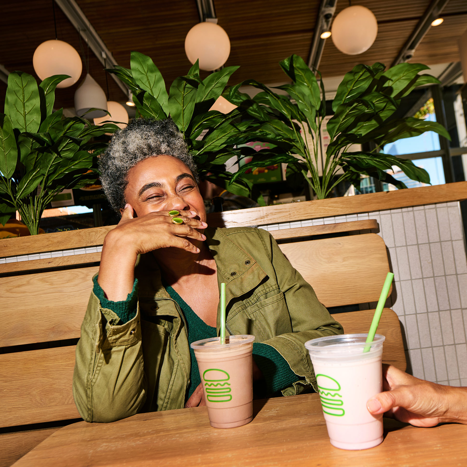 A person with short curly hair laughs while sitting at a wooden booth in a restaurant, holding a chocolate milkshake. Another hand offers a pink milkshake. Green plants and hanging lights are in the background.