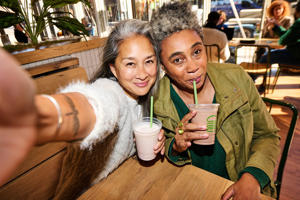 Two women sit closely together in a café, smiling and taking a selfie. They each hold a cup with a straw, appearing to enjoy milkshakes. Natural light fills the cozy space with plants and wooden furniture.