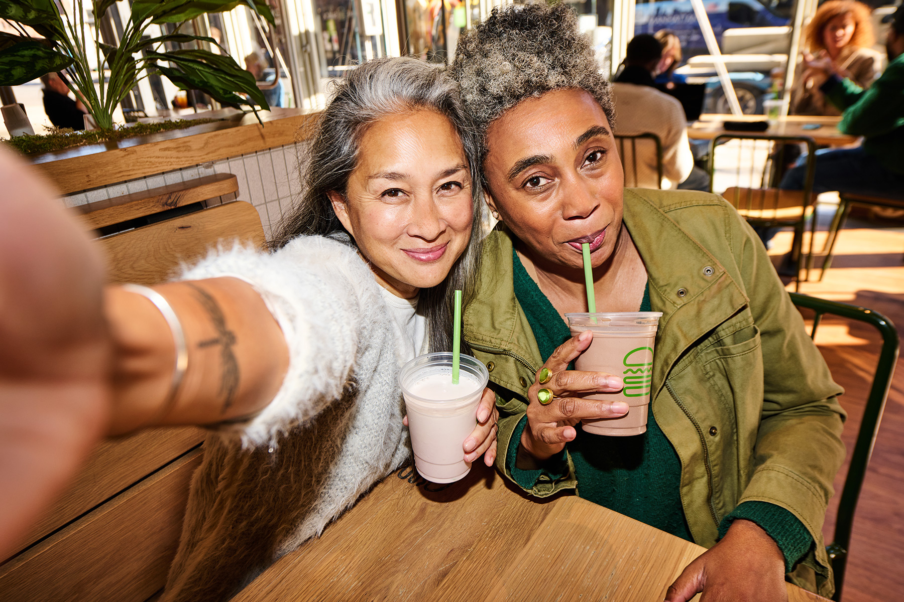 Two women sit closely together in a café, smiling and taking a selfie. They each hold a cup with a straw, appearing to enjoy milkshakes. Natural light fills the cozy space with plants and wooden furniture.