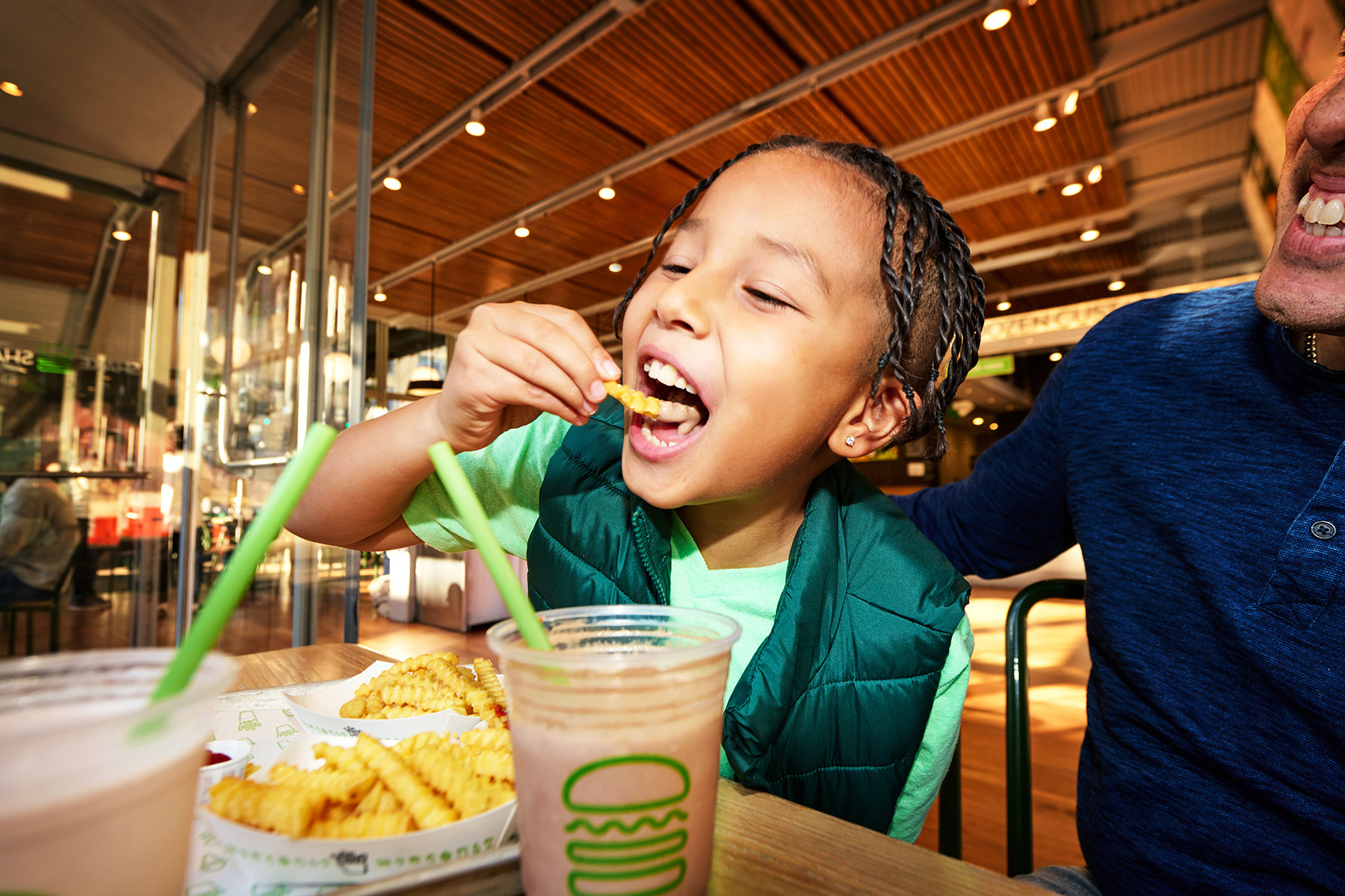 A child with braided hair and a green vest eats crinkle-cut fries at a restaurant, smiling. Two shakes with green straws and trays of fries are on the table. An adult in a blue shirt sits beside the child.