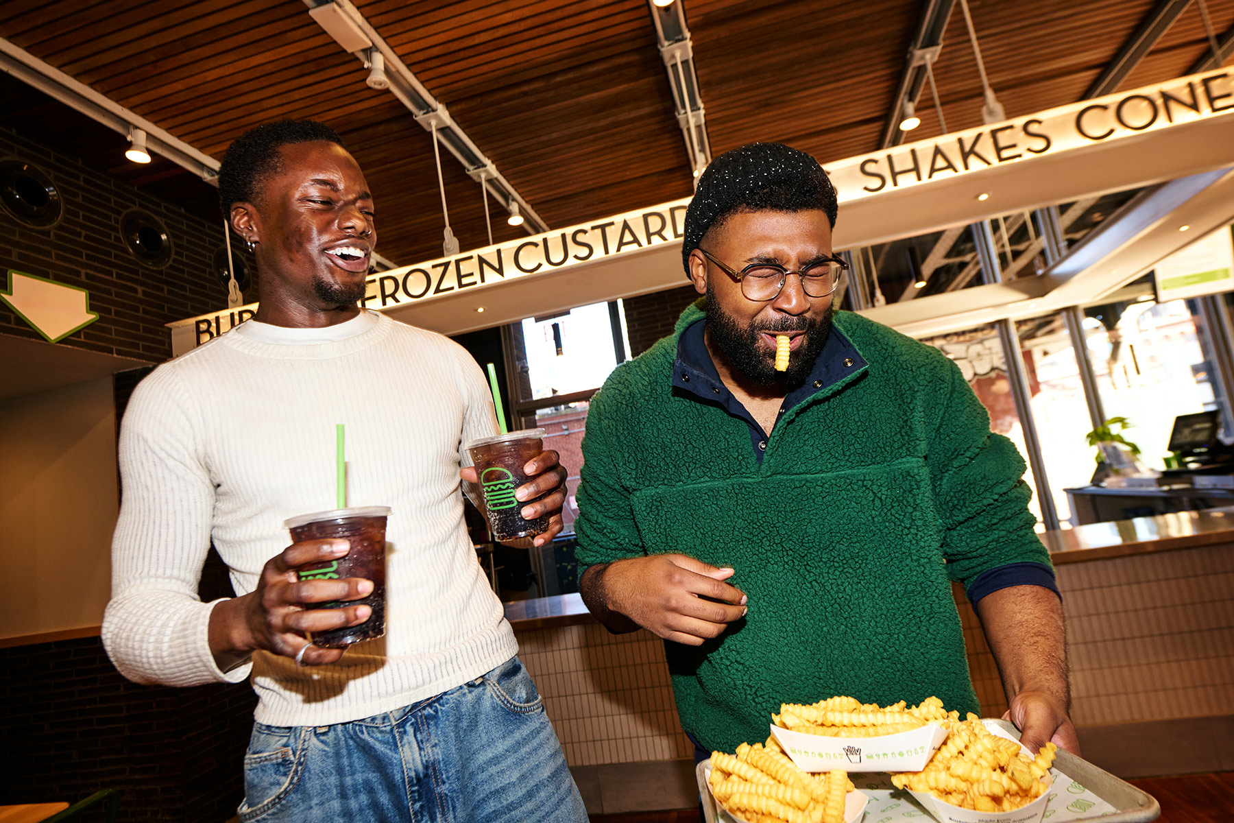 Two men laugh together inside a restaurant; one holds two drinks, while the other, with fries in his mouth, holds a tray of crinkle-cut fries. A menu sign above reads “FROZEN CUSTARD,” “SHAKES,” and “CONES.”.