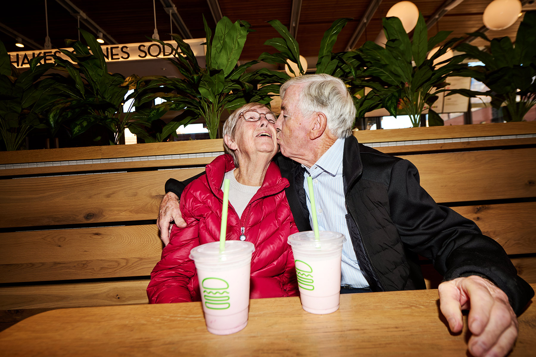 An elderly man kisses an elderly woman on the cheek as they sit together in a booth, smiling and holding two pink milkshakes in front of them. Lush green plants and a wooden wall are in the background.