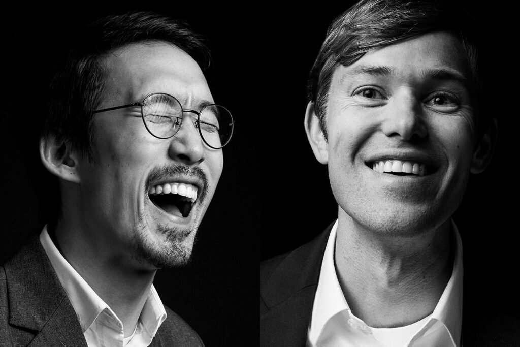 Black and white photo of two men in suits, each smiling. The man on the left is laughing with his eyes closed and wearing round glasses. The man on the right is smiling, looking directly ahead. Both are shown from the shoulders up.