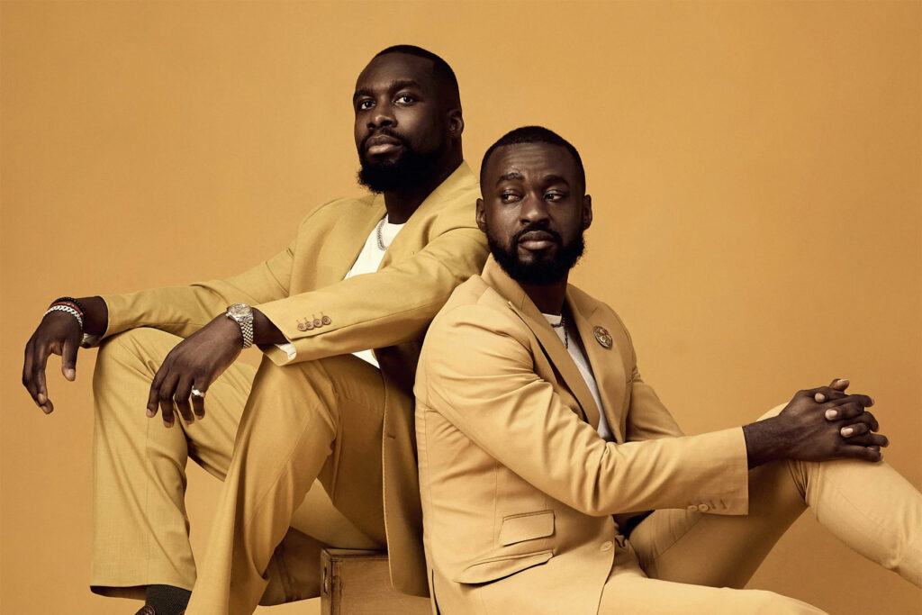 Two men with beards wearing matching light tan suits and white shirts pose against a solid tan background. One sits on a crate with his arm resting on his knee; the other sits on the floor, both looking off in different directions.